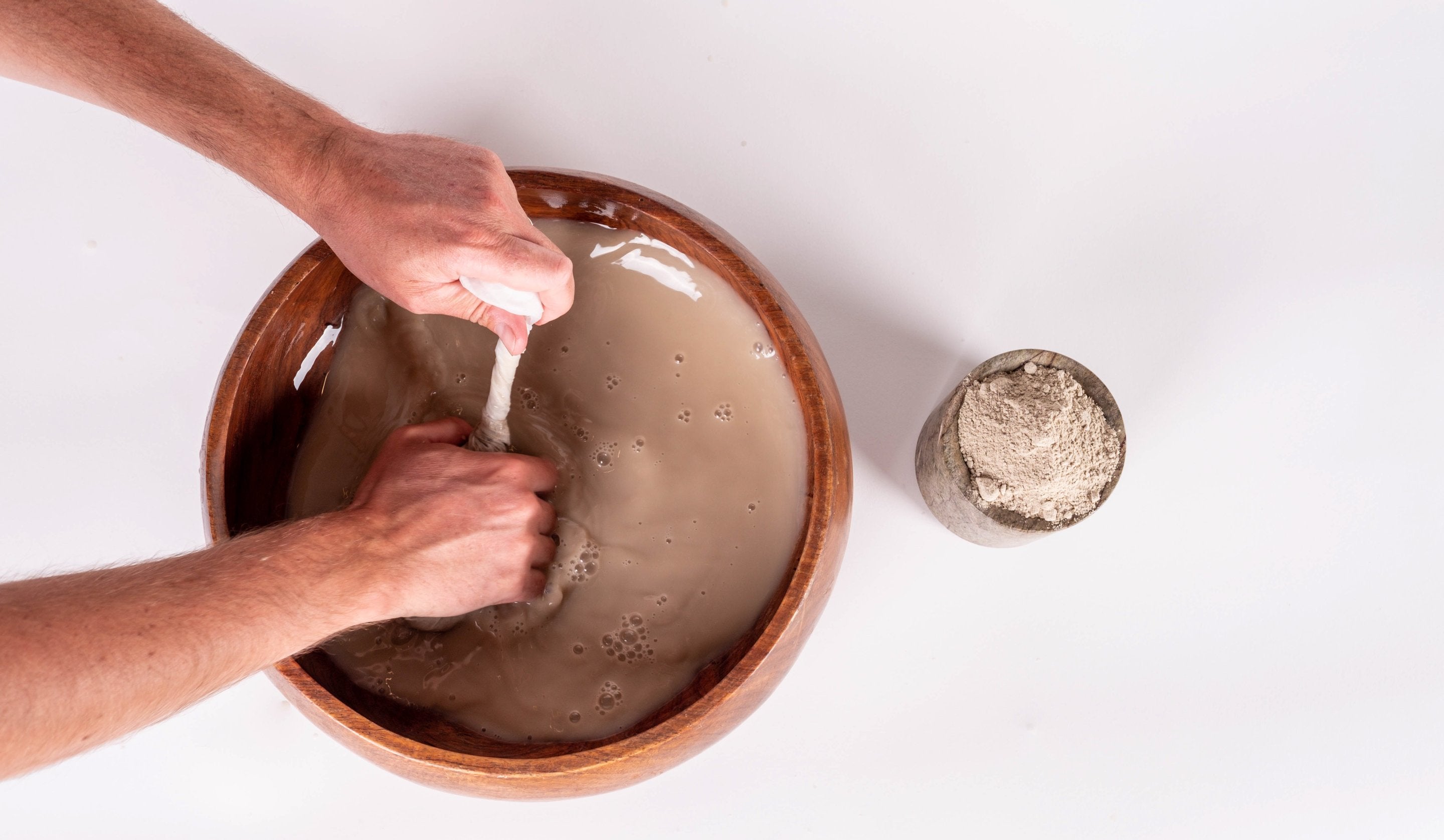 Hands kneading kava in a strainer bag in a wooden kava bowl, beside a small cup of ground kava root powder